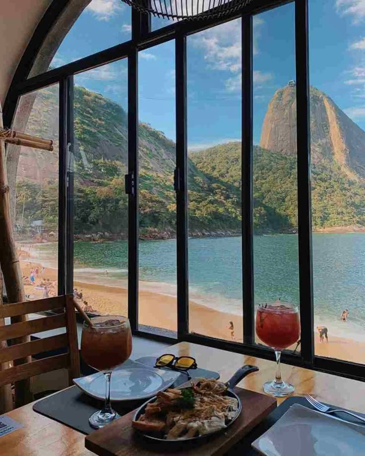 Dining with ocean view: scenic beach, mountains, and delicious meal at a window-side table. Refreshing drinks and stunning landscape.