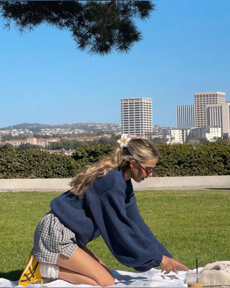 Young woman enjoys a sunny picnic, kneeling on a blanket in a park, wearing a navy sweater and checked skirt, city skyline backdrop.
