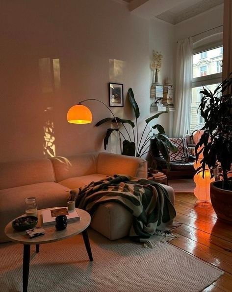 Cozy living room with a beige sofa, warm lighting, plants, and a soft blanket. Calm ambiance for relaxing or reading.
