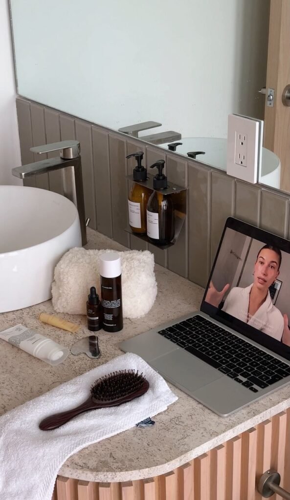 Aesthetic Skincare Routine Bathroom vanity with skincare products, laptop playing a video, towel, brush, and a mirror reflecting bottles, creating a cozy routine space. - Handy Home Bathroom vanity with skincare products, laptop playing a video, towel, brush, and a mirror reflecting bottles, creating a cozy routine space.