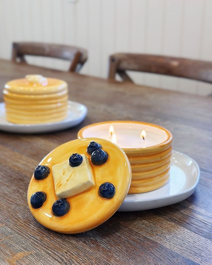 Unique pancake-themed candle with burning wicks on a wooden table; lid features butter and blueberry design. Cozy home décor.