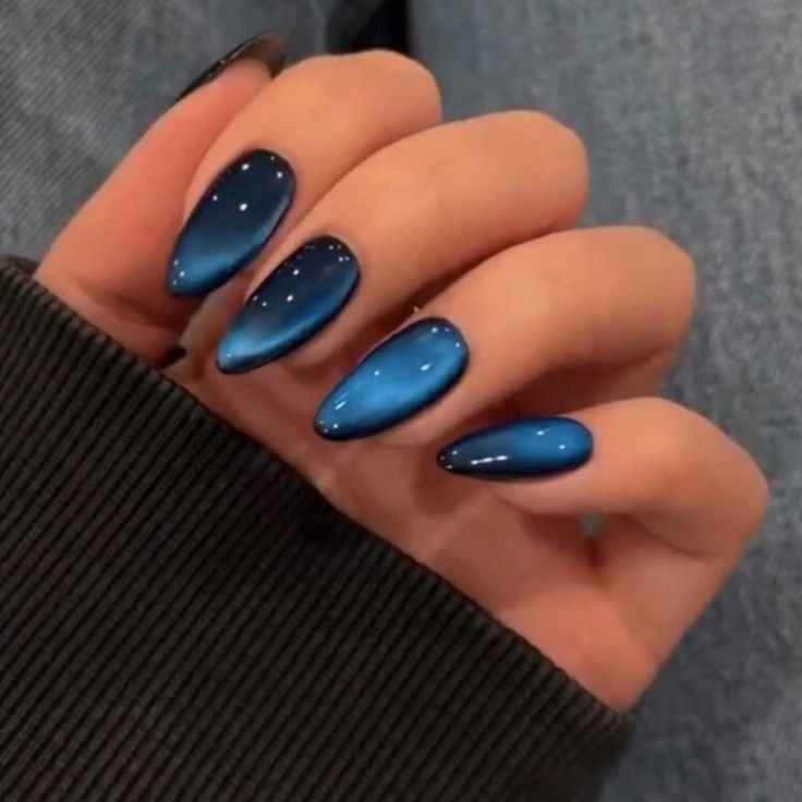 Blue Press On Nails _ Color_ Blue _ Size_ Os Close-up of hand with blue metallic almond-shaped nails, wearing a dark sweater, showcasing elegant nail art design. - Handy Home Close-up of hand with blue metallic almond-shaped nails, wearing a dark sweater, showcasing elegant nail art design.