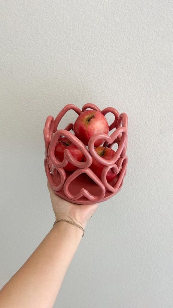 Hand holding a pink ceramic basket filled with red apples against a white wall background.