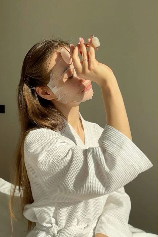 Woman in a robe applies a face mask and uses a face roller, enjoying a skincare routine in natural sunlight.