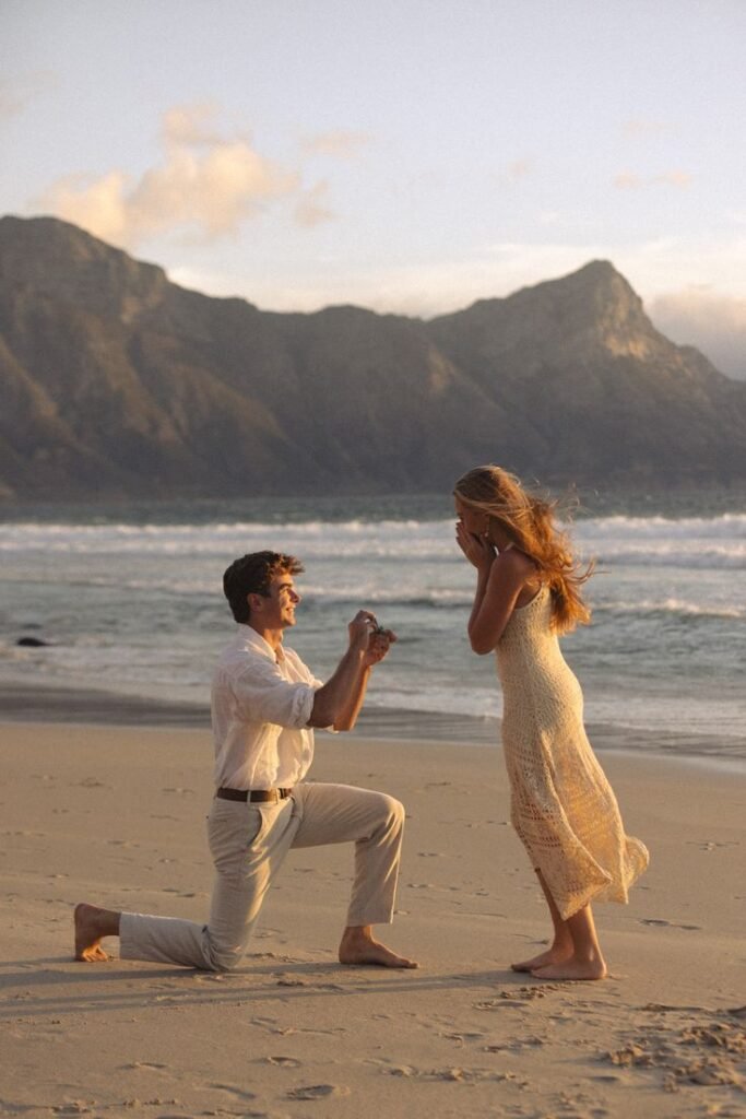 Man proposing on a beach at sunset, woman surprised and happy, mountains in background. Romantic engagement moment.