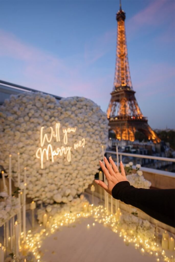 Romantic proposal by the Eiffel Tower with a heart of white roses and Will you marry me? lights at sunset.