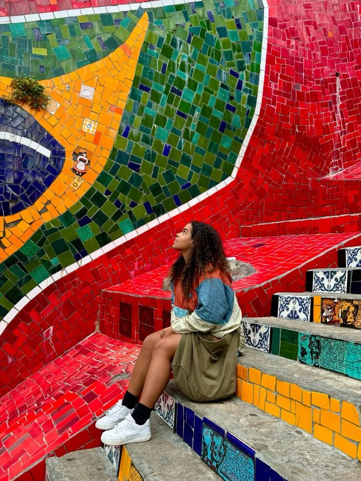 Woman sitting on colorful mosaic steps, gazing upwards at vibrant tiled wall. Artistic urban scene filled with bright colors.