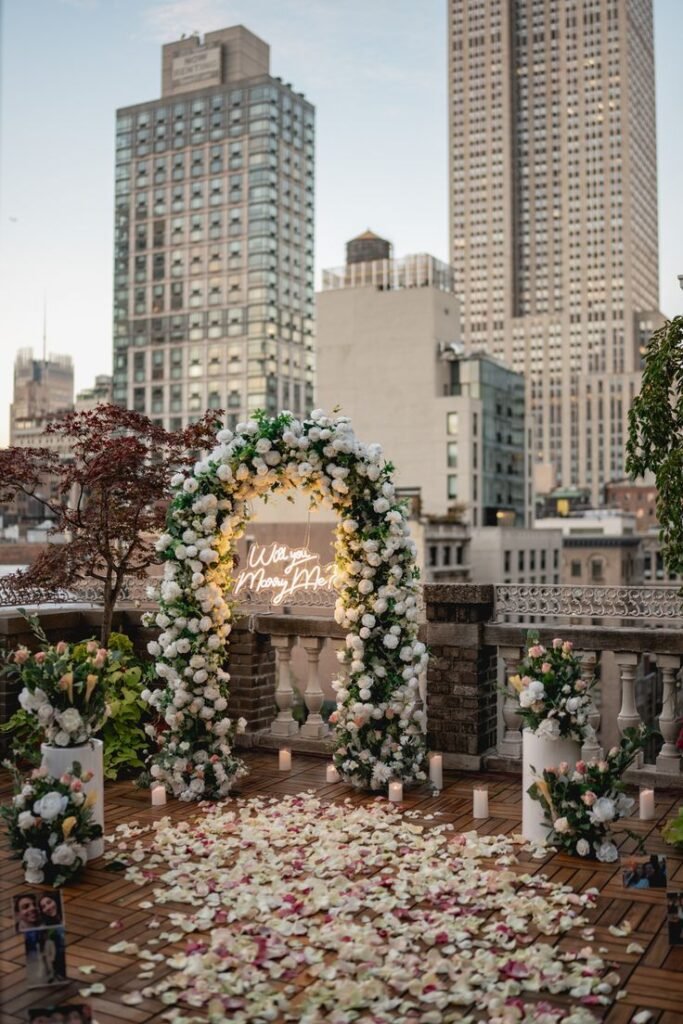 Romantic rooftop proposal setup with floral arch, Will you marry me? sign, candles, and rose petals against city skyline backdrop.