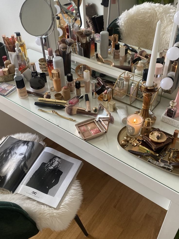Vanity table with assorted makeup, brushes, candle, and open fashion magazine on a cozy chair.