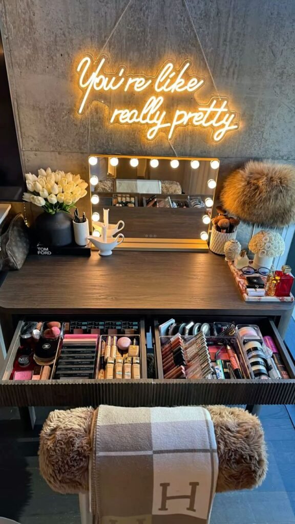 Makeup vanity with illuminated mirror, organized cosmetics, and You're like really pretty neon sign on textured wall.