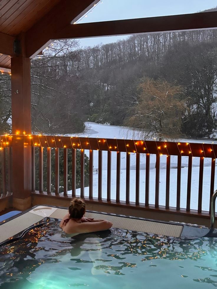 Person relaxing in a hot tub with snowy forest view and twinkling lights for a cozy winter retreat.