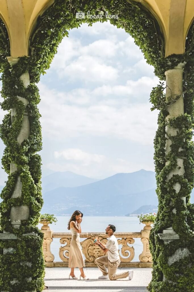 Marriage proposal on a picturesque balcony overlooking a serene lake and mountains, framed by lush greenery.
