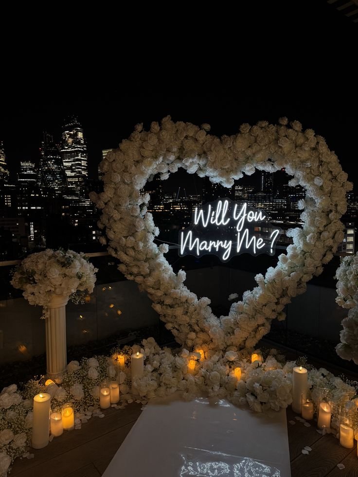 Romantic city rooftop proposal with Will You Marry Me? in neon inside a heart of white roses, surrounded by candles. Marriage Proposal Ideas