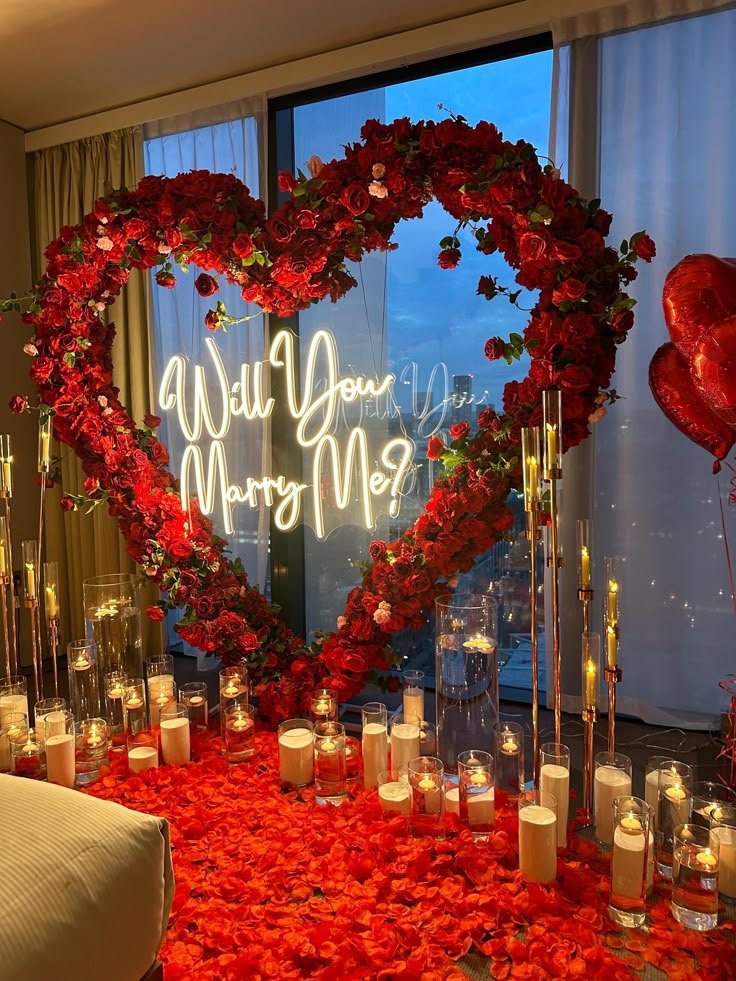 Romantic proposal setup with heart-shaped rose arch and Will You Marry Me? sign, surrounded by candles and petals.