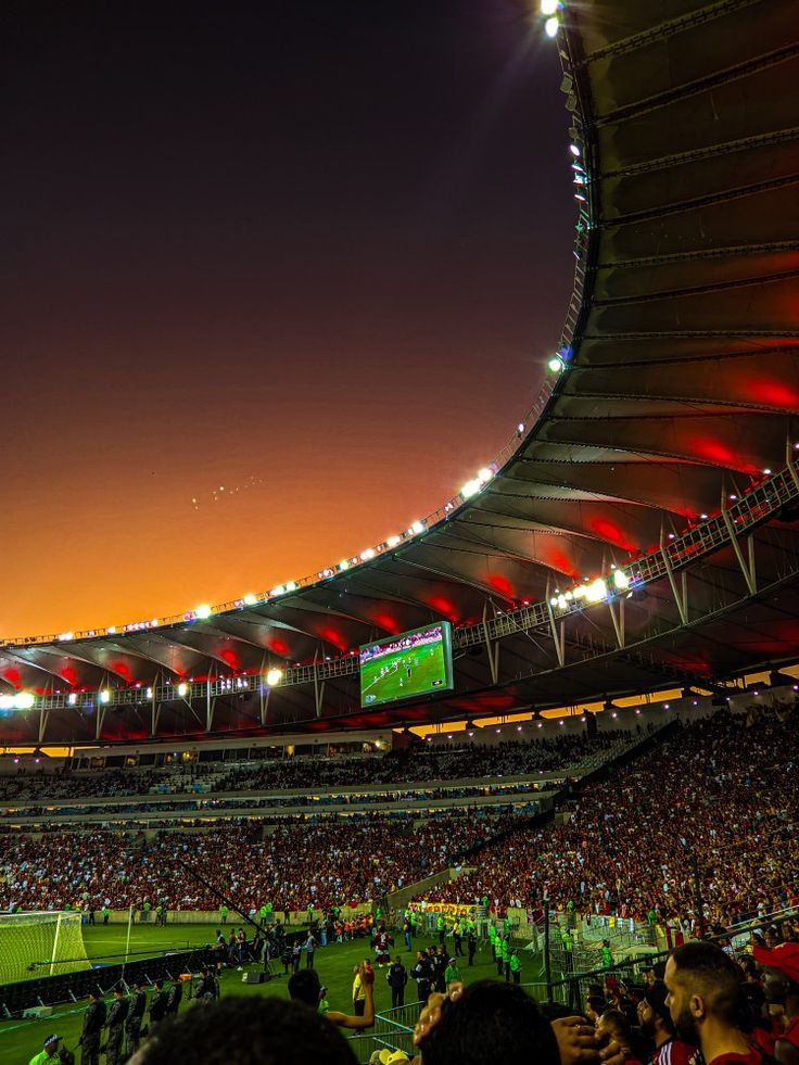 Vibrant sunset over a packed soccer stadium with illuminated stands and fans cheering during an exciting match, creating a lively atmosphere.