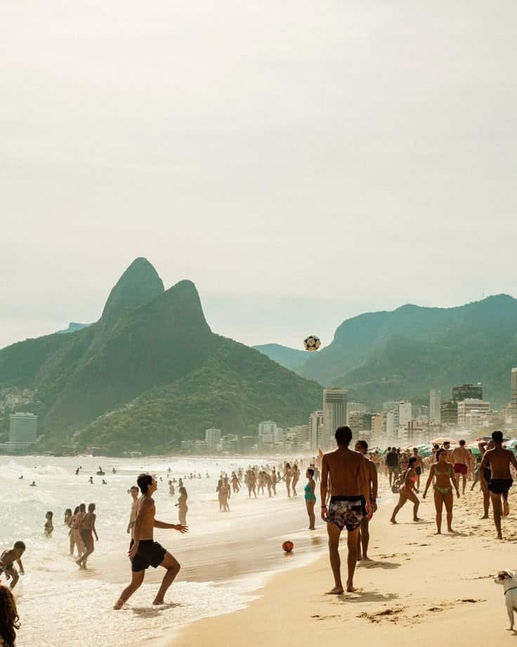 Crowded beach with people playing soccer under the sun, scenic mountains in the background. Perfect day at Ipanema Beach, Rio.