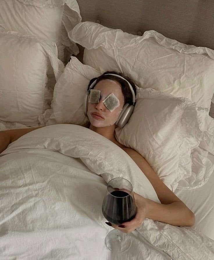 PAMPER __ PINK Woman relaxing in bed with face mask, headphones, tea bags on eyes, holding a glass of red wine. Spa day relaxation mood. - Handy Home Woman relaxing in bed with face mask, headphones, tea bags on eyes, holding a glass of red wine. Spa day relaxation mood.