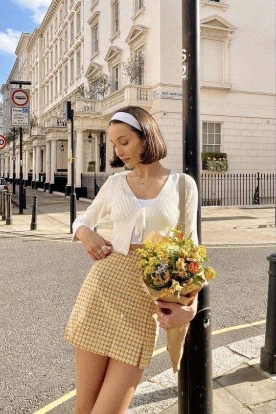 Woman in white cardigan holding flowers, standing on a sunny street in an urban setting, stylish outfit, city architecture.