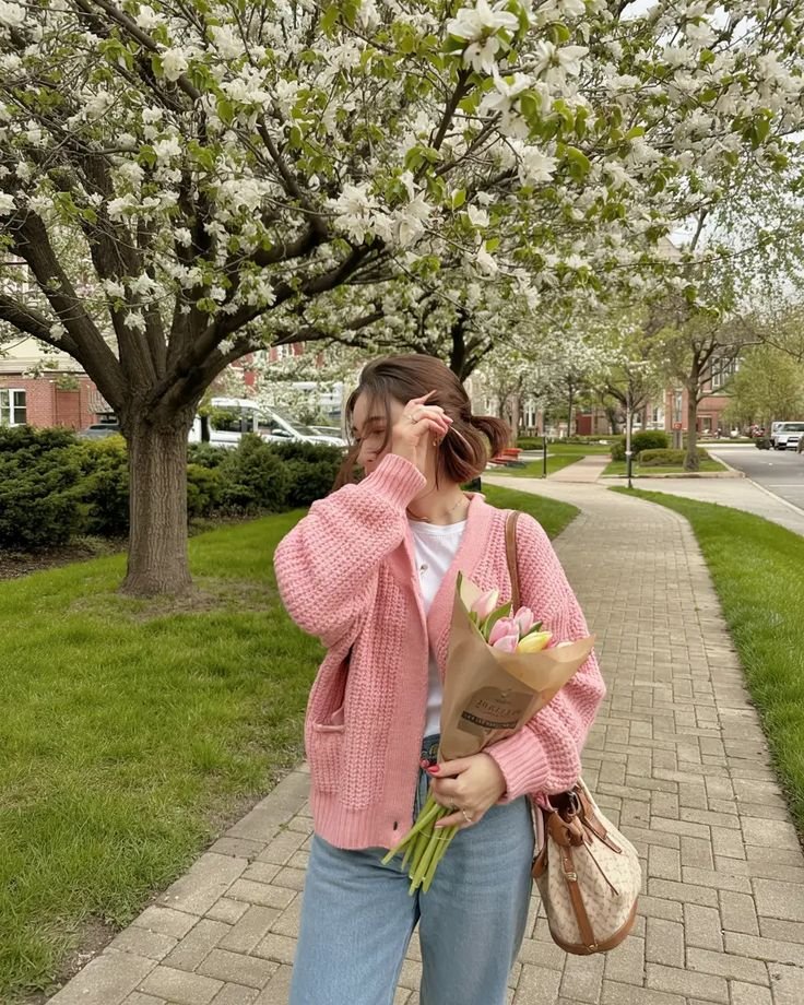 Woman in pink cardigan holds tulips under blooming trees on a sunny day, enjoying spring vibes and a leisurely walk.