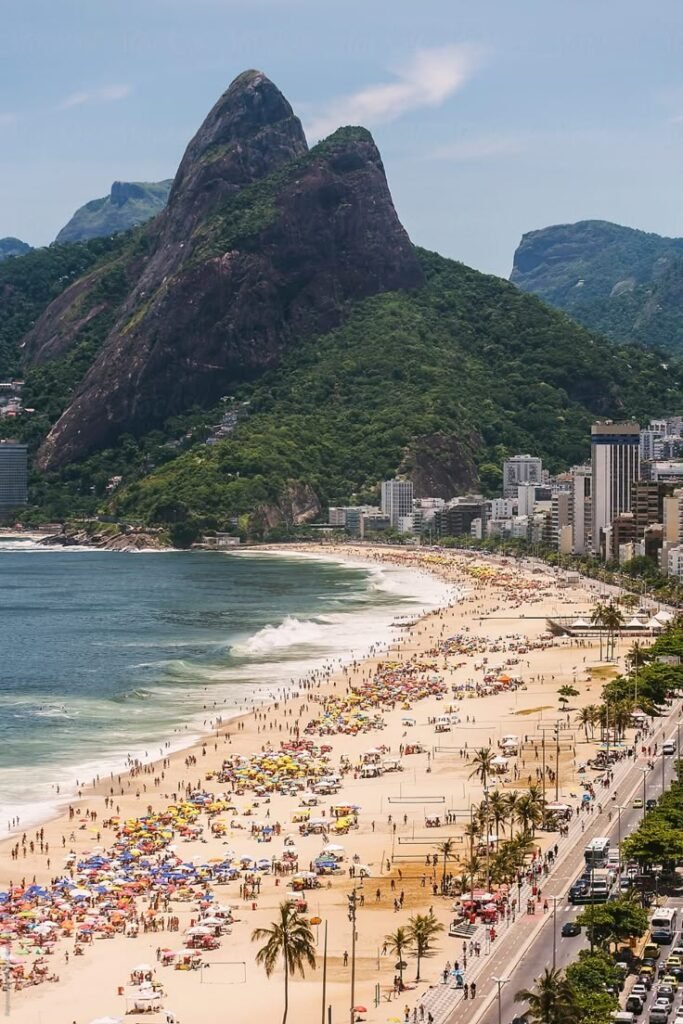 Sunny Ipanema Beach with crowds, colorful umbrellas, and view of Dois Irmãos mountains in Rio de Janeiro, Brazil.