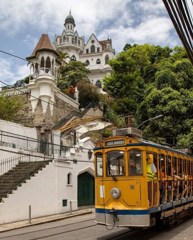 Yellow tram passes a historic, castle-like building in lush, tree-lined street under a partly cloudy sky.