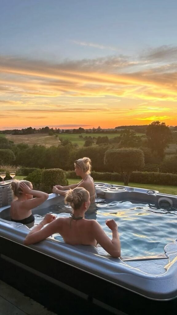 Three people relaxing in a hot tub, enjoying a scenic sunset view over lush green fields. Perfect for a peaceful getaway.