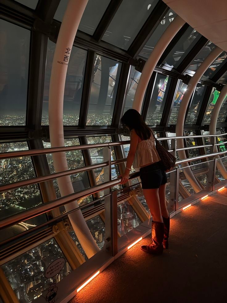 Person gazing at cityscape from a tower observation deck at night, surrounded by illuminated city lights and urban skyline.