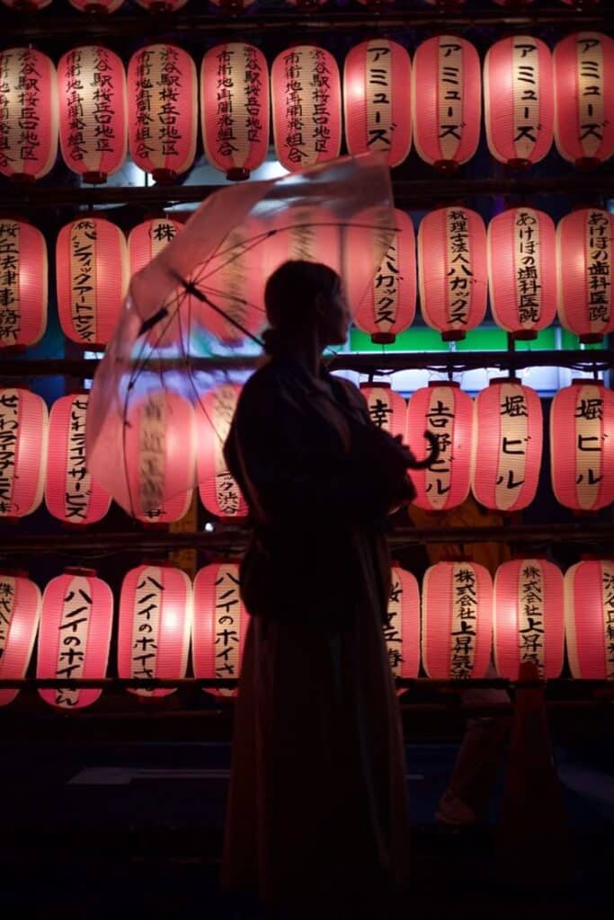 Silhouette of a person with umbrella against a backdrop of glowing Japanese lanterns at night.