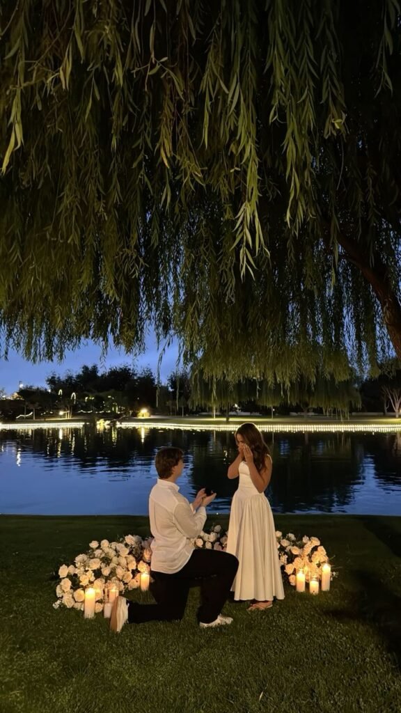 Romantic outdoor proposal by a lake with candles and flowers, couple sharing a special moment under a tree at dusk.