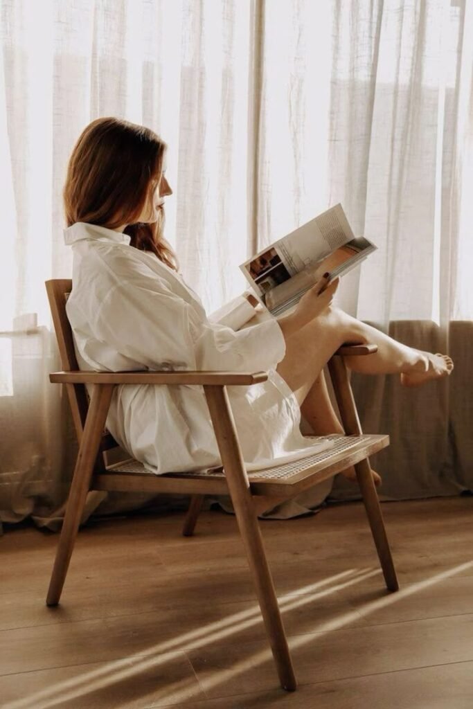 afa0d0a6-966e-4528-9a8d-60b706145c26 Woman in white shirt reads a magazine on a wooden chair, sunlight streaming through sheer curtains, creating a cozy atmosphere. - Handy Home Woman in white shirt reads a magazine on a wooden chair, sunlight streaming through sheer curtains, creating a cozy atmosphere.