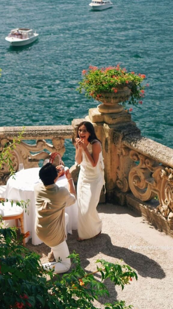 Romantic proposal by the lake; joyful woman in white dress reacts to ring, surrounded by scenic views and boats.