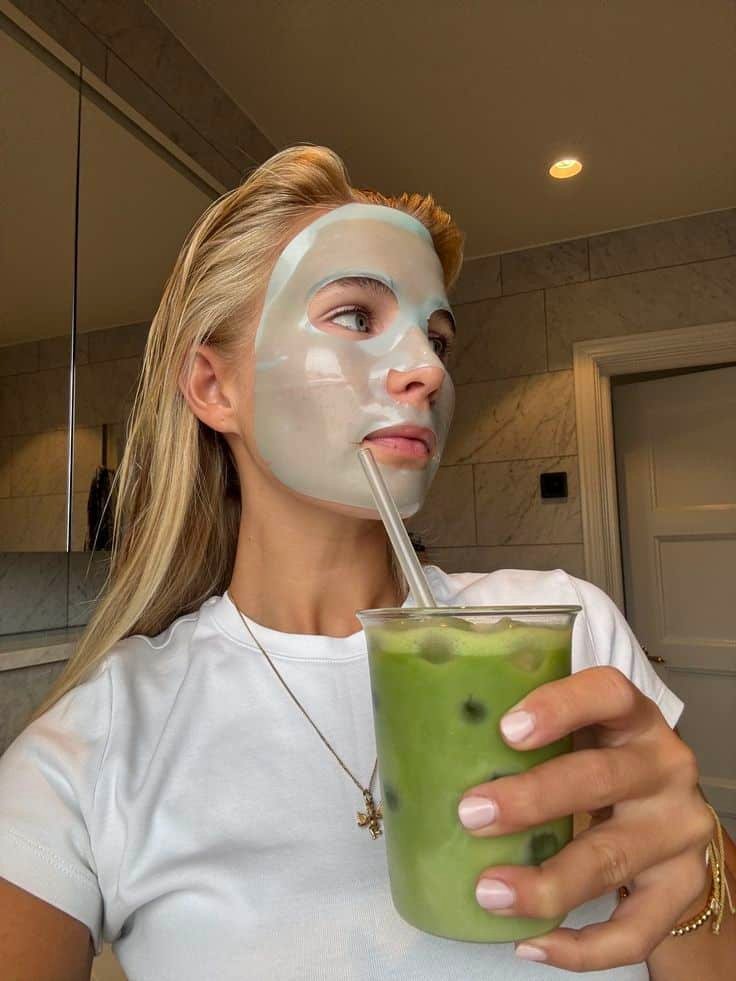 Woman with a face mask drinks iced matcha latte in a stylish bathroom, capturing a relaxing skincare and wellness moment.
