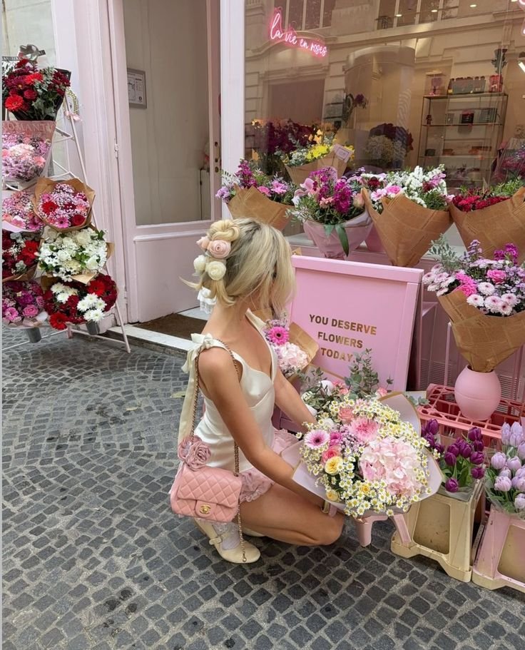 Woman with floral hairstyle admires colorful bouquets outside a pink flower shop, capturing a charming Parisian vibe.