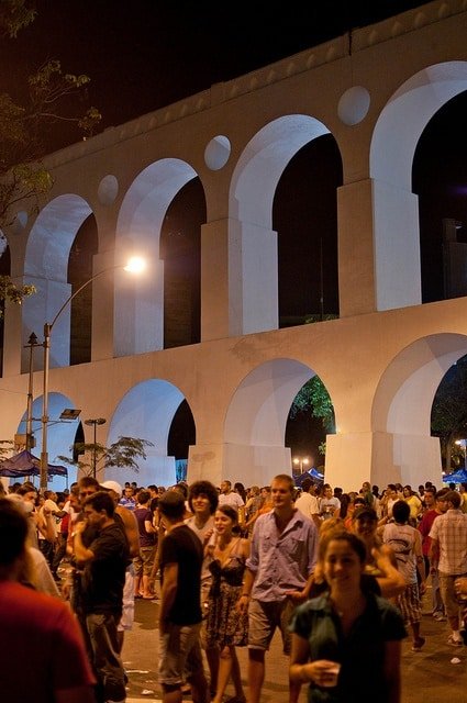 A lively crowd enjoys a vibrant night under the illuminated arches of Lapa, Rio de Janeiro, with the iconic aqueduct in view.