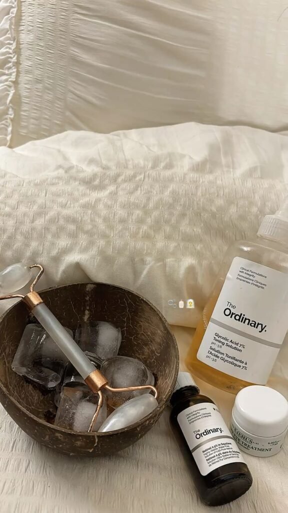 Skincare essentials: jade roller in a bowl with ice, The Ordinary and Kiehl’s products on a cozy white pillow background.