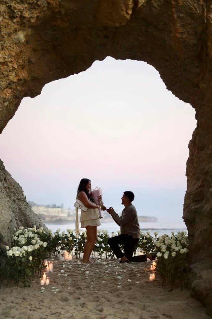 Beach proposal at sunset in scenic cave setting, surrounded by flowers and candles. Romantic and unforgettable moment.
