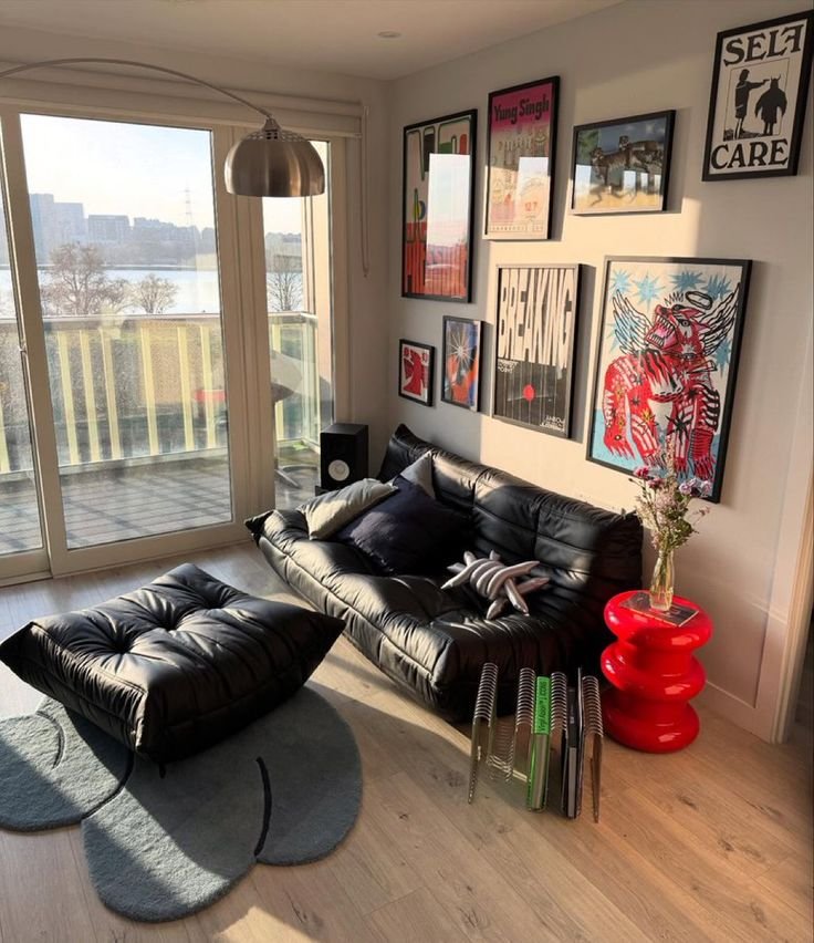 Modern living room with black leather sofa, colorful art, and a skylit balcony view. Cozy design with eclectic decor accents.