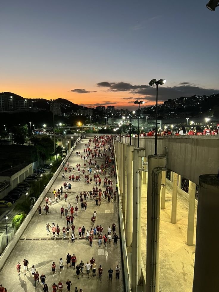 Crowd of people in red shirts leaving a stadium walkway at dusk, with a cityscape and colorful sunset in the background.