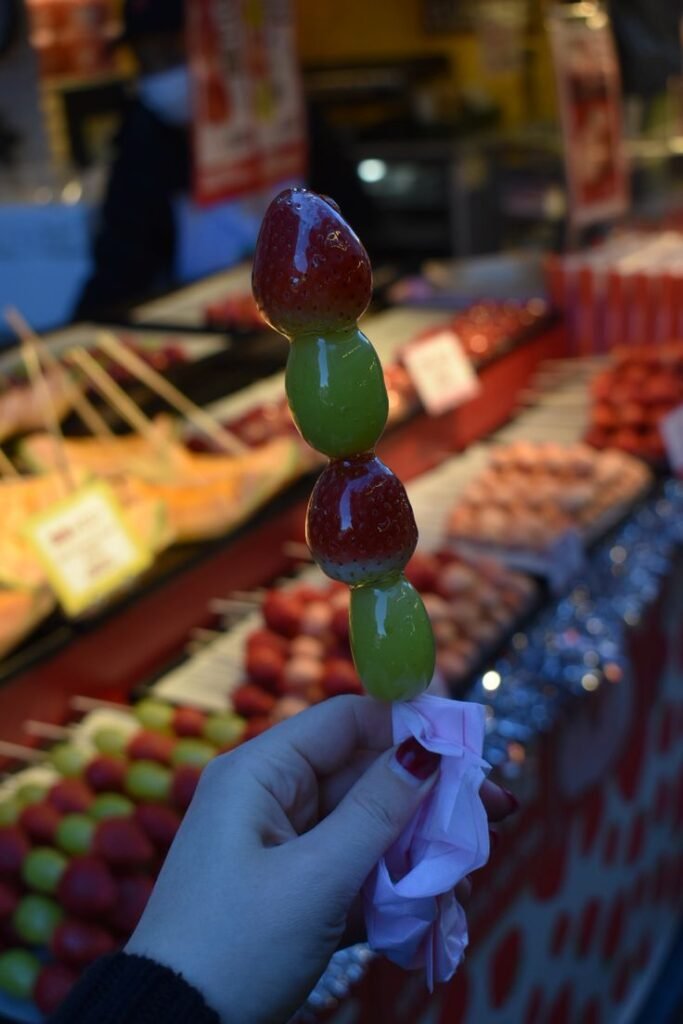 Candied fruit skewer held at a vibrant market stall, featuring glazed strawberries and grapes. Perfect street food treat.