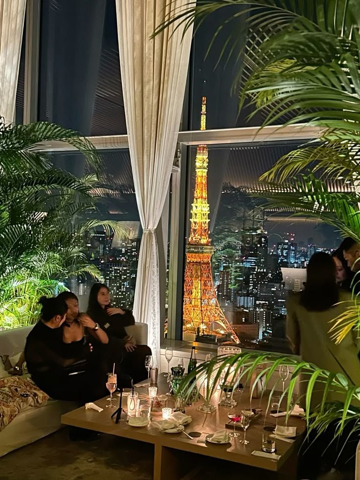 Cozy lounge with people enjoying a night view of Tokyo Tower through large windows, surrounded by lush indoor plants.