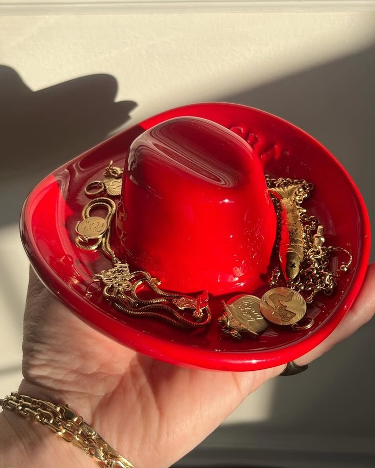 Red cowboy hat bowl with gold jewelry and coins, held in a hand, illuminated by sunlight. Perfect for country-themed decor. Useful Clay Crafts