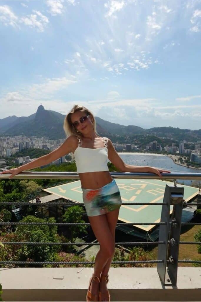 Woman in stylish outfit poses with scenic cityscape and mountains in the background, capturing a sunny day on a balcony.