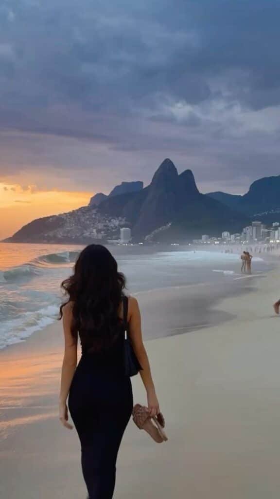 Woman in black dress walking on Ipanema beach at sunset with Dois Irmãos mountains in the background.