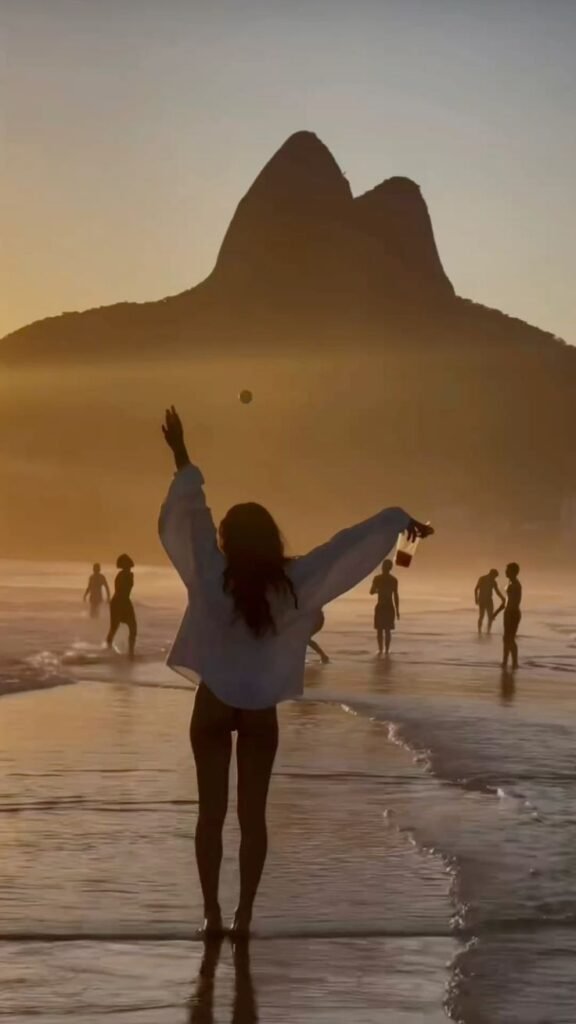 Person enjoys sunset on a vibrant beach with distant mountains and beachgoers, capturing carefree summer vibes.