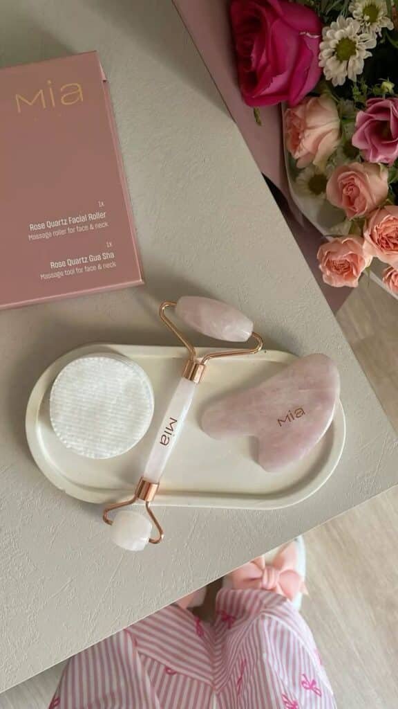 Rose quartz facial roller and gua sha on a tray, with flowers in the background. Perfect for skincare and relaxation rituals.