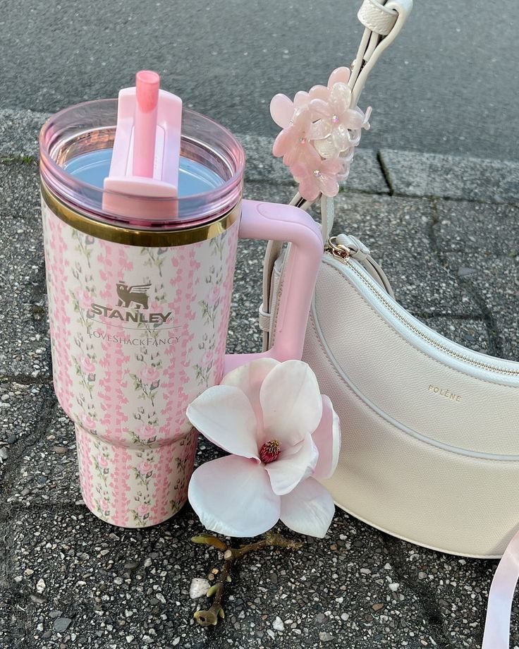 Pink floral Stanley cup with straw beside a white Polene bag and magnolia flower on pavement. Stylish spring accessories.