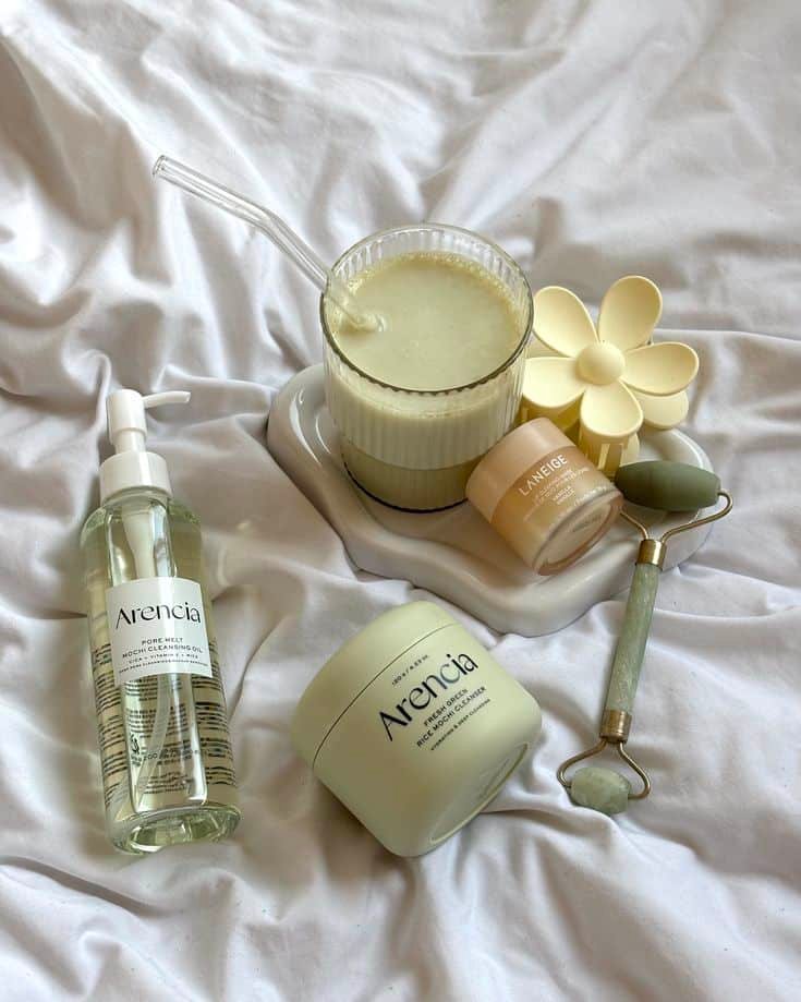Skincare essentials on white sheets: Arencia products, jade roller, cream jar, and smoothie for a refreshing self-care routine.